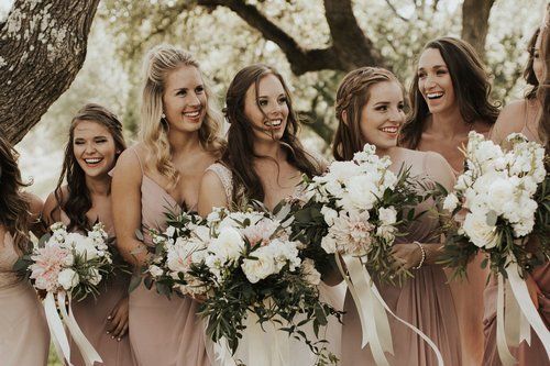 Five women in blush dresses holding white floral bouquets at an outdoor wedding