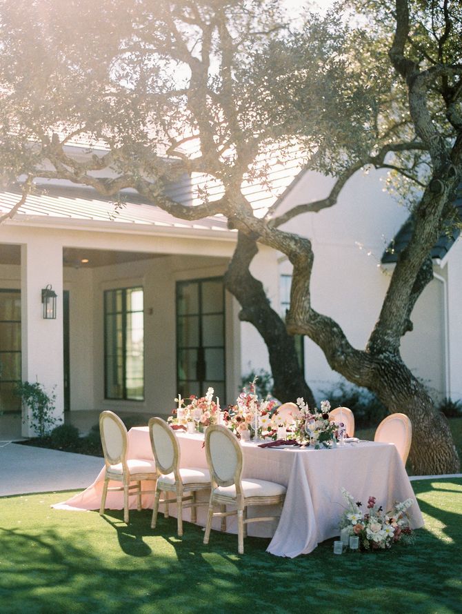 Outdoor dining table set with white chairs and floral centerpiece under trees beside a house
