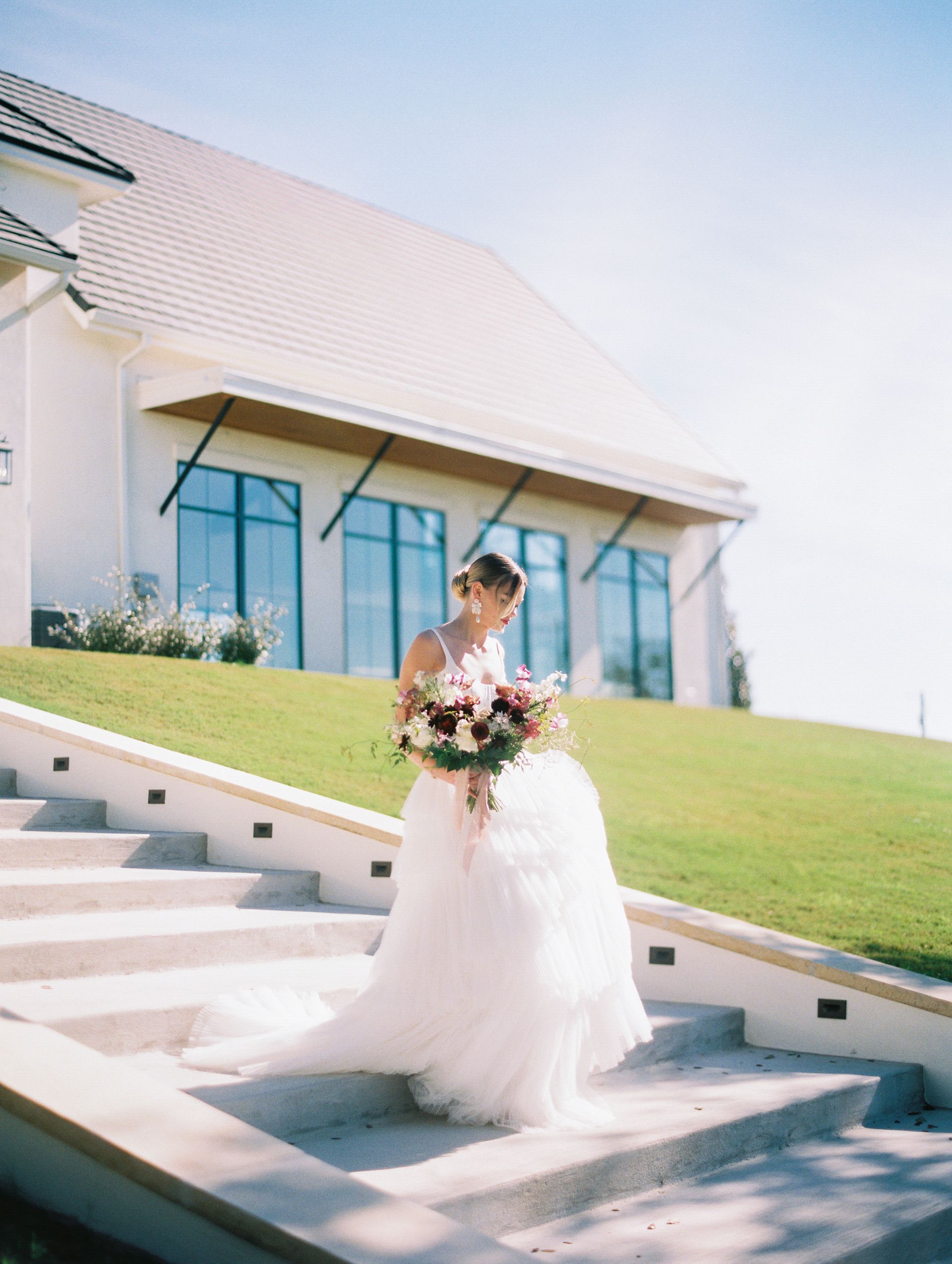 Bride in a white gown holding a bouquet on outdoor steps beside a modern white building