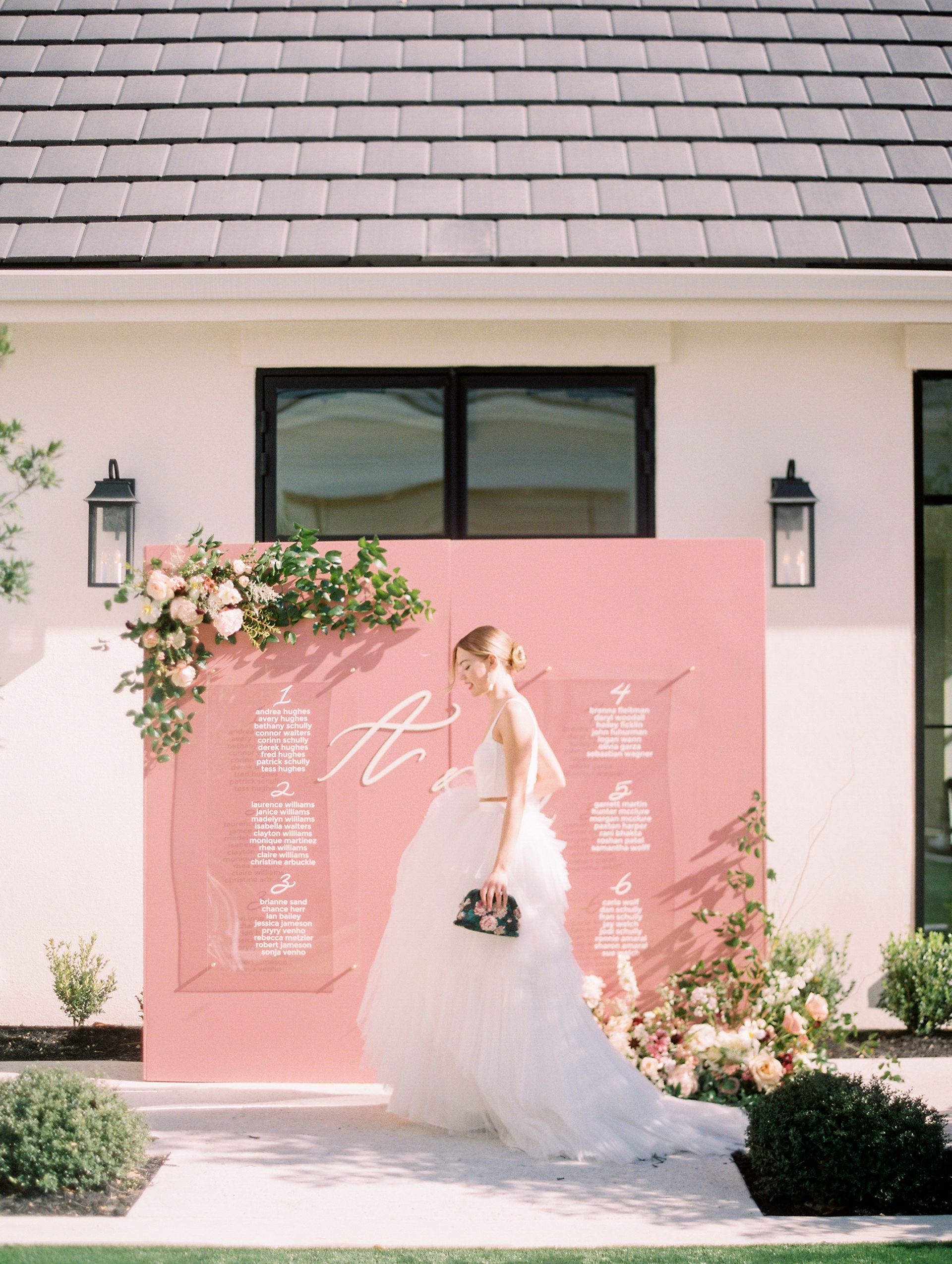 Bride in white gown posing before a pink floral wedding backdrop outside a building