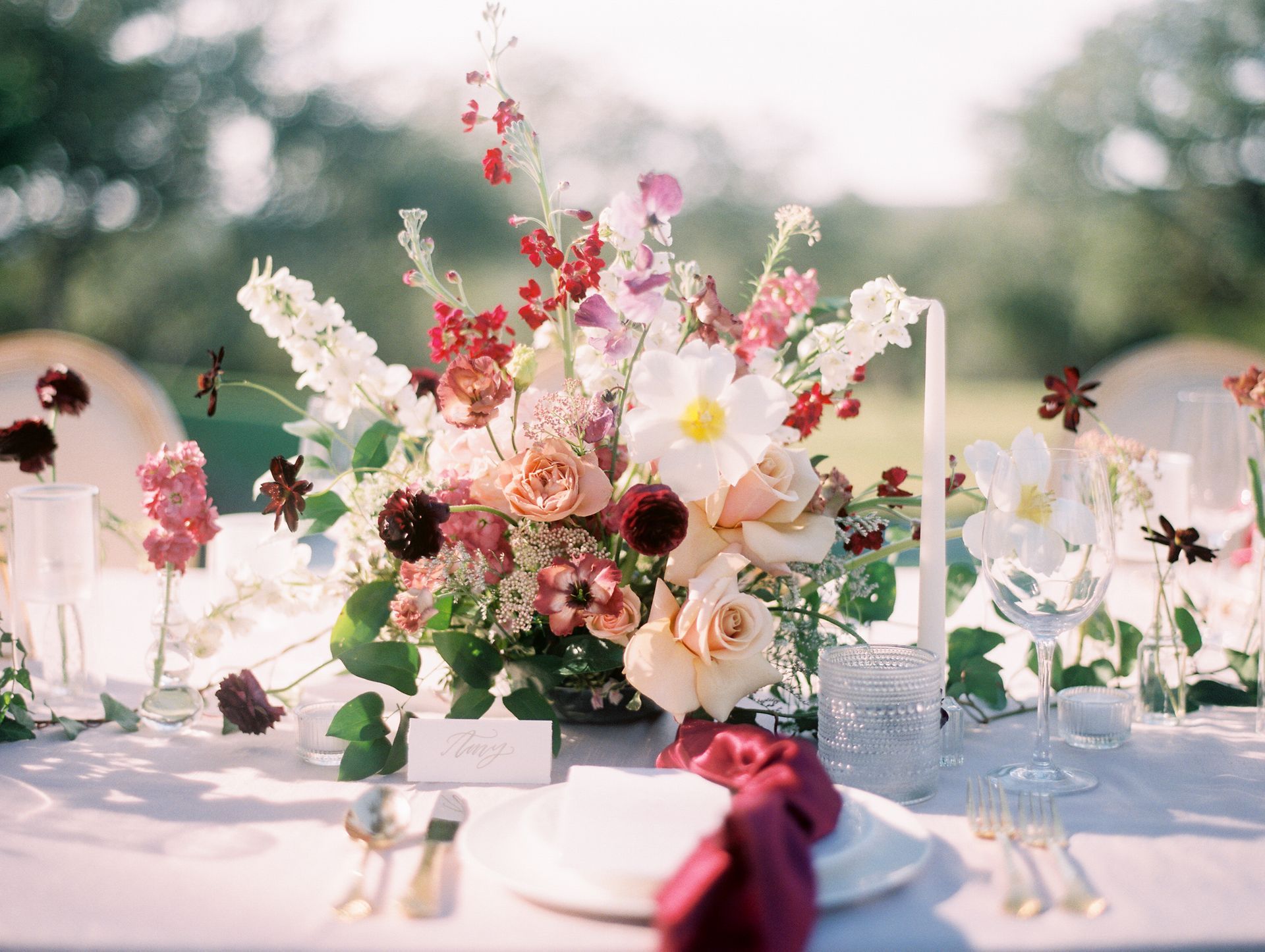 Elegant outdoor table setting with pink and white floral centerpiece, glassware, and burgundy napkins on a white tablecloth