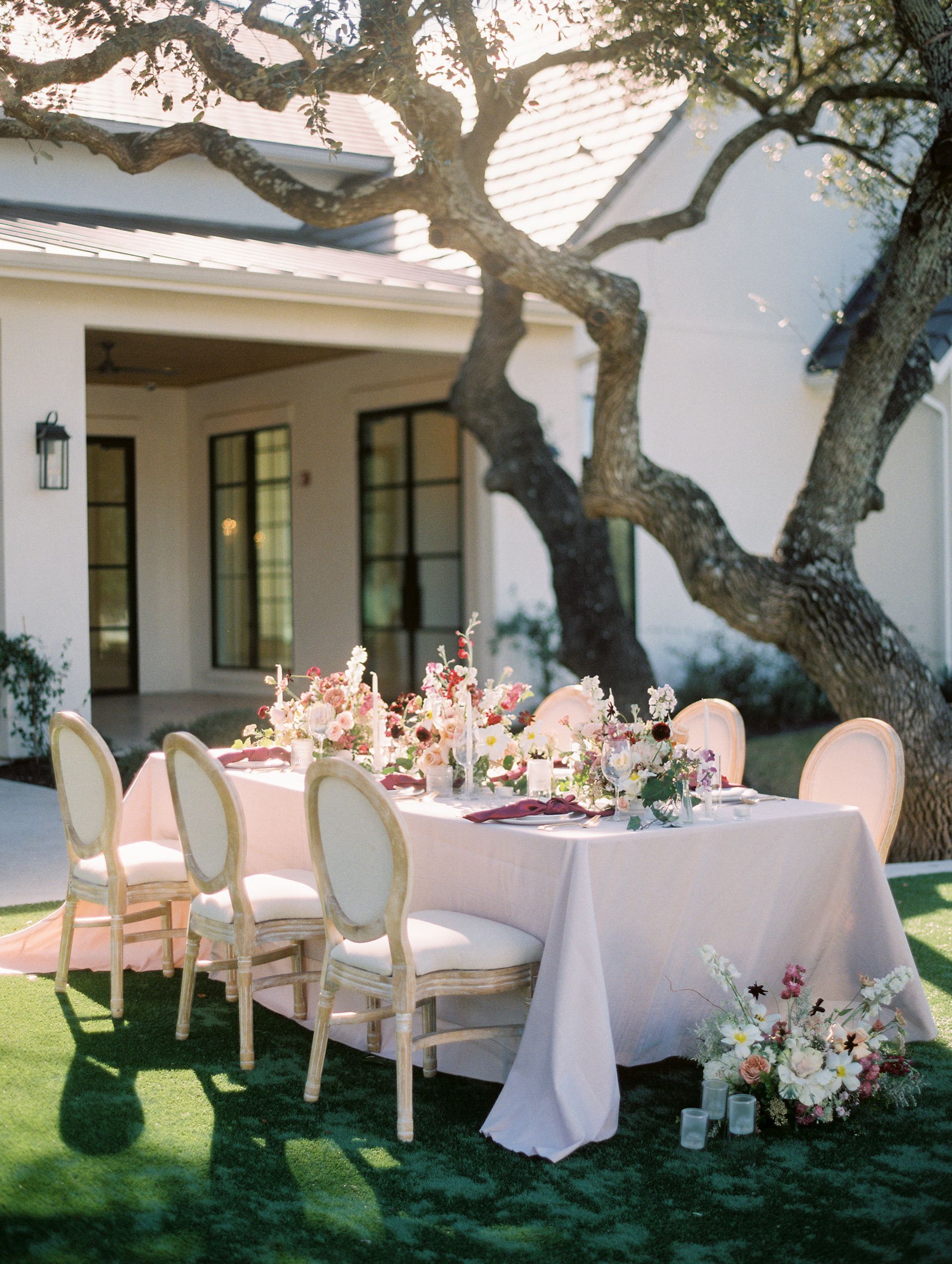 Elegant outdoor dining table set under a large tree beside a white stucco house.