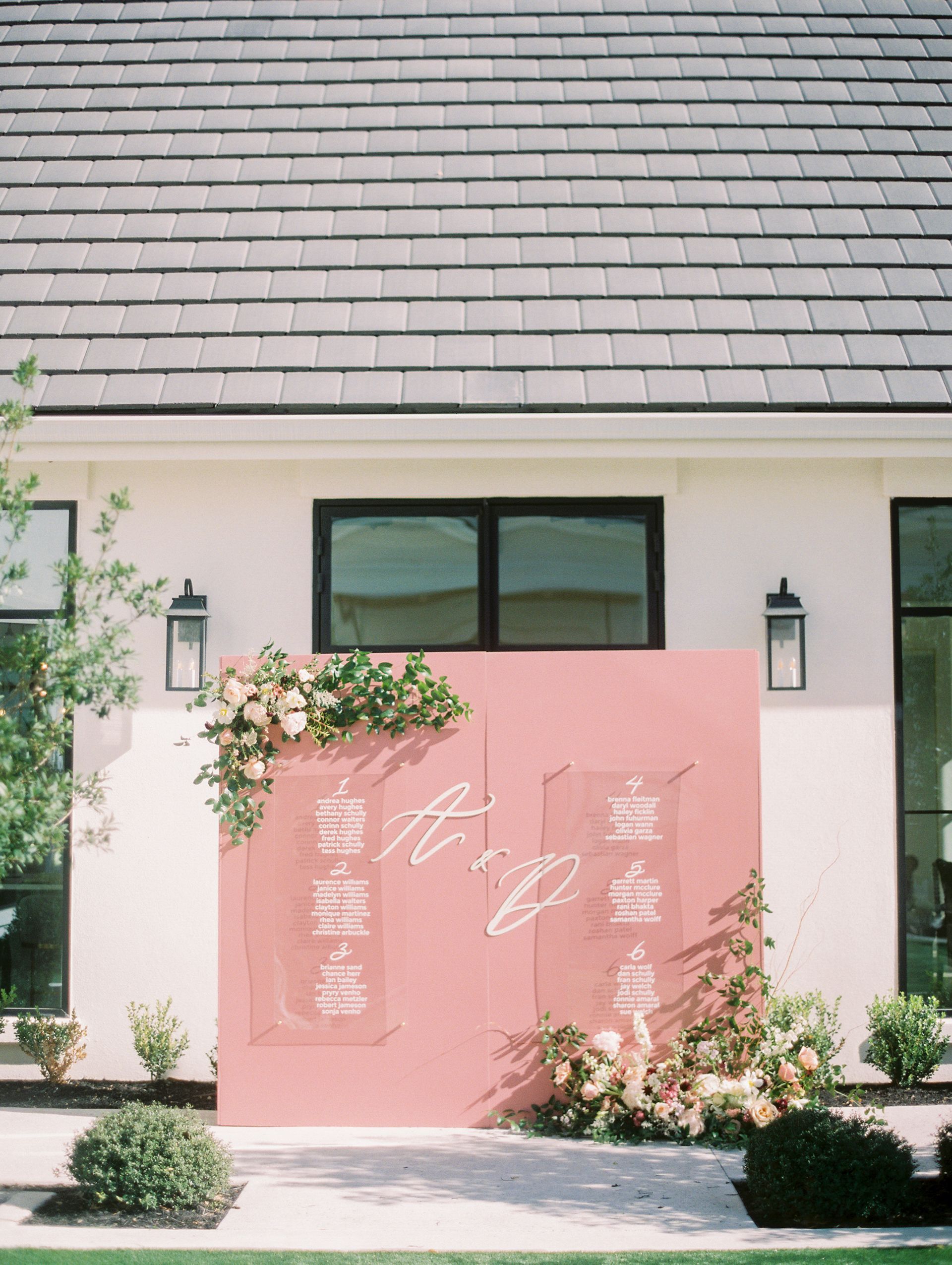 Pink welcome sign with white floral text outside a house, framed by greenery and lanterns