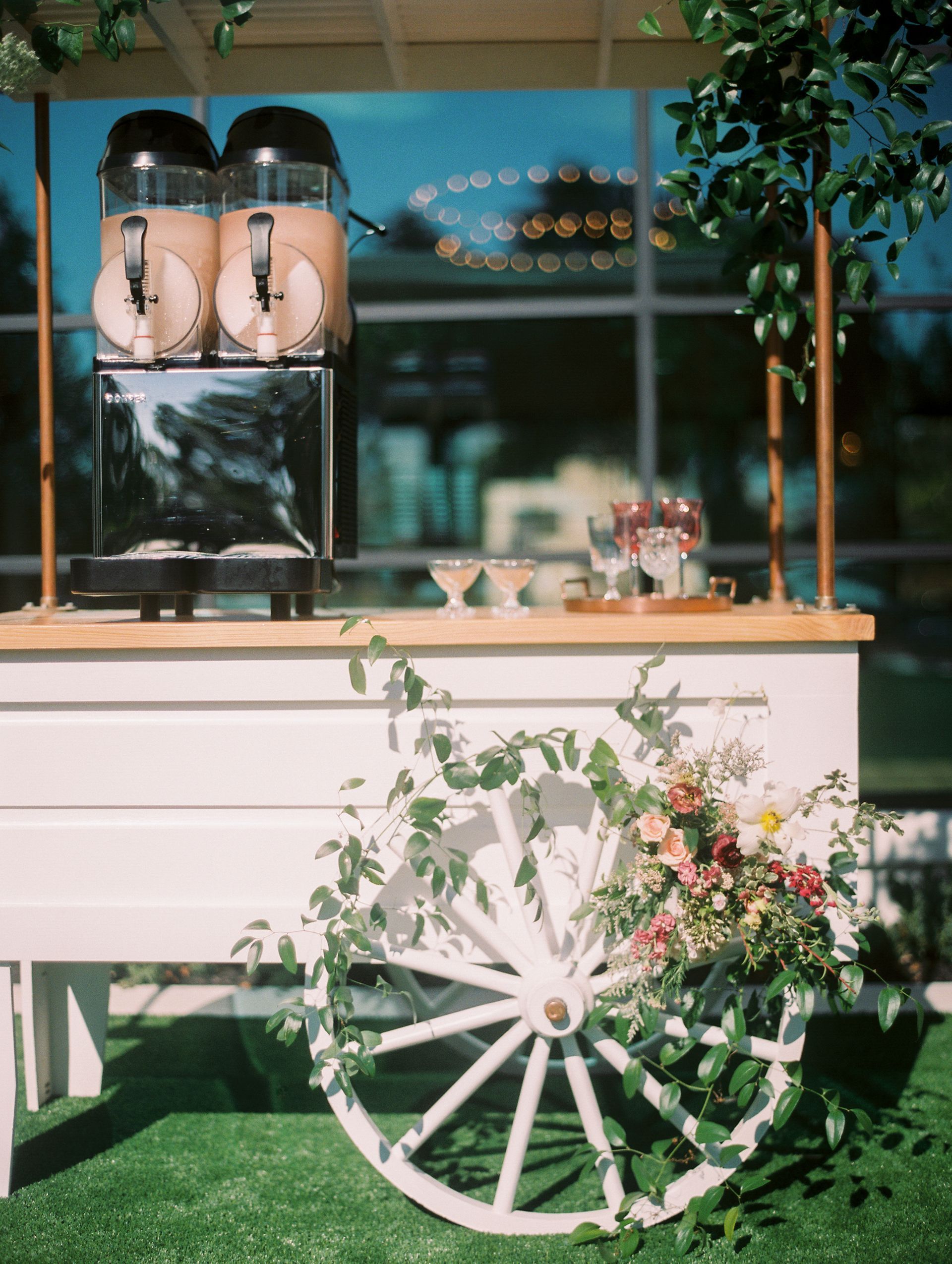 Outdoor coffee cart with white wheel decor, flowers, and glass drink dispensers under a wooden awning.