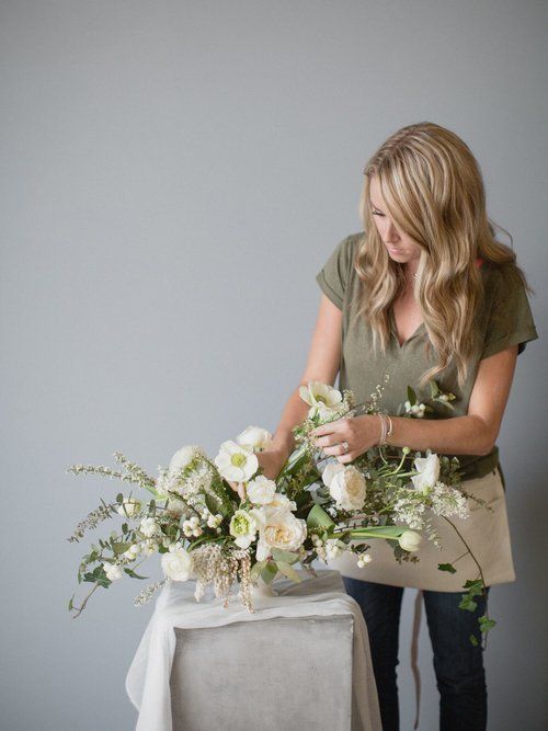 Woman arranging white floral centerpiece on a table against a gray wall.