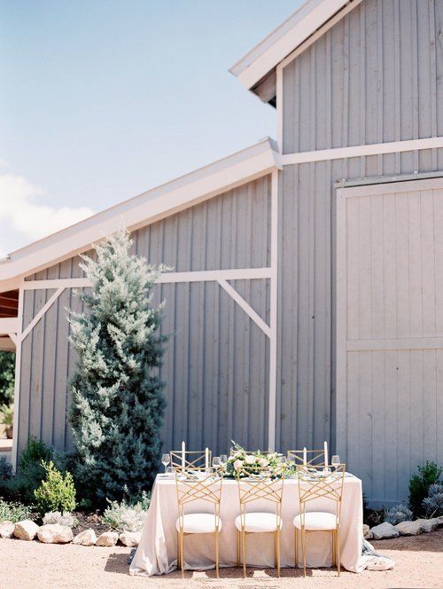 Outdoor table set with white chairs beside a gray barn-style building and small trees