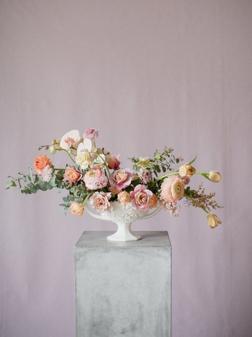 Pastel floral arrangement in a white vase on a gray pedestal against a mauve backdrop