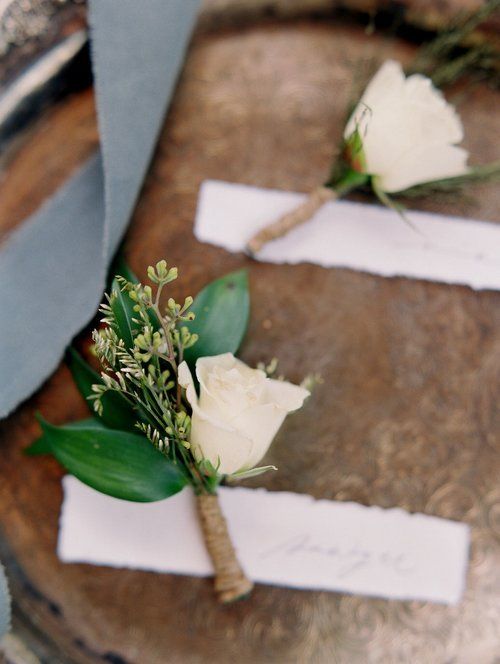 White floral boutonnieres with green leaves attached to ribbon tags on a rustic surface