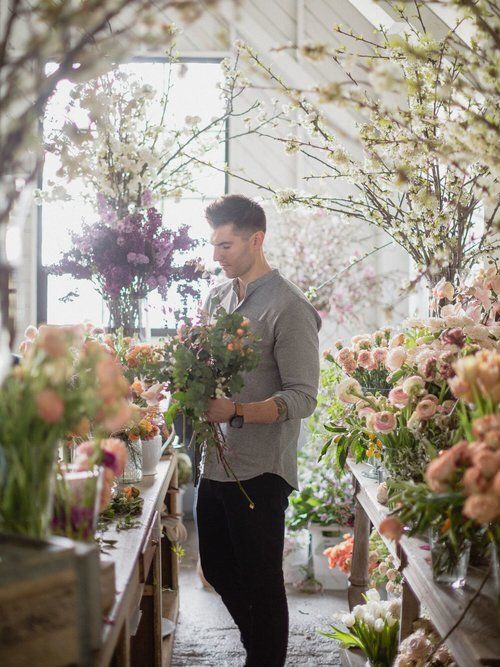 Person arranging flowers in a bright floral shop aisle, surrounded by bouquets and greenery.
