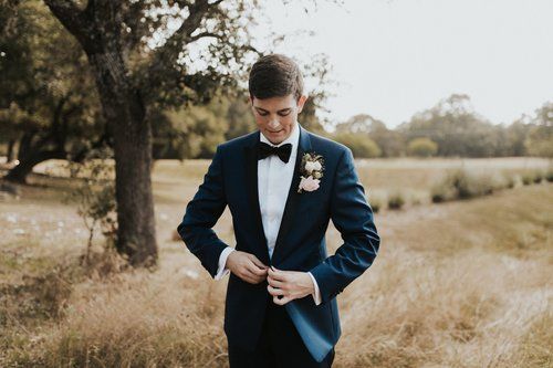 Man in a navy suit adjusting his jacket outdoors in a grassy field near trees