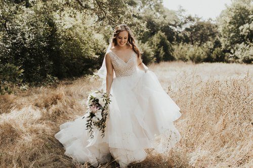 Bride in a white gown walking through a sunlit grassy field with trees in the background