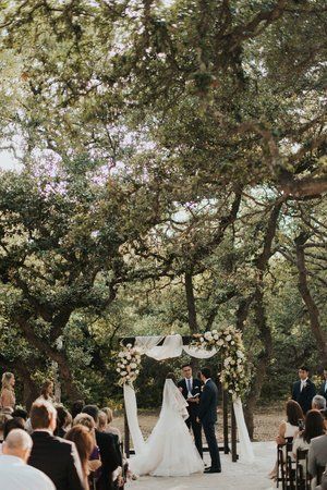 Outdoor wedding ceremony beneath large trees, with bride and groom at the altar and guests seated along the aisle