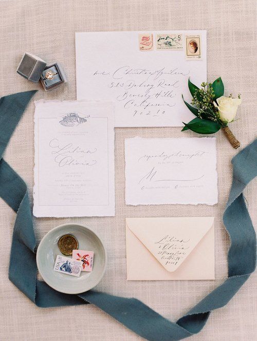 Elegant wedding stationery set with white envelopes, calligraphy, stamps, flowers, and blue ribbon on a pale background