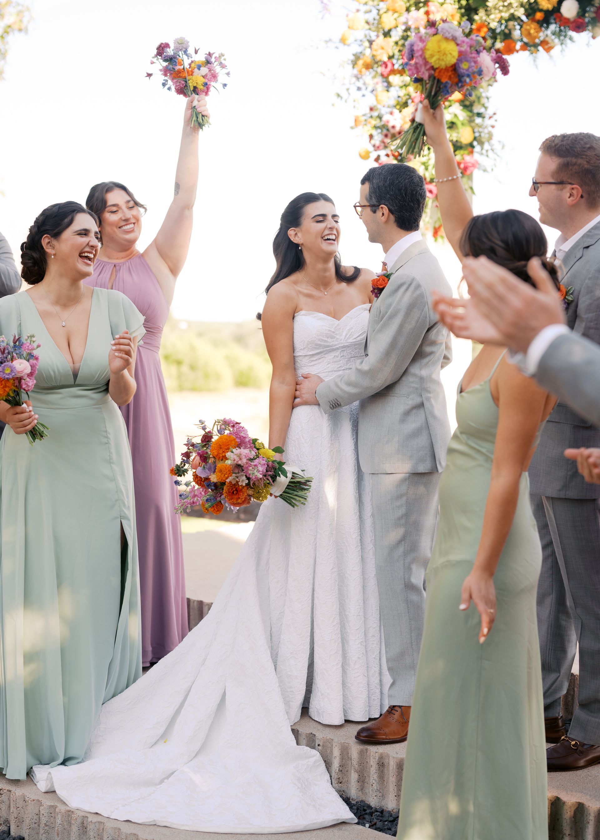 Wedding couple kissing under floral arch, bridesmaids cheering nearby outdoors