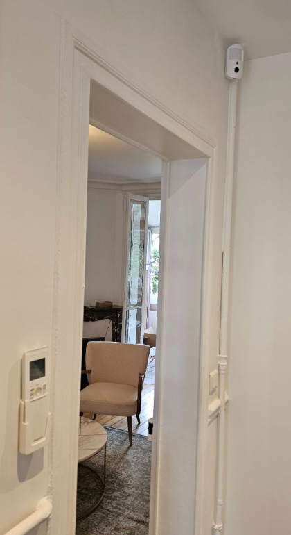 Une porte blanche révèle un coin salon meublé. Une caméra de sécurité et un thermostat sont visibles.