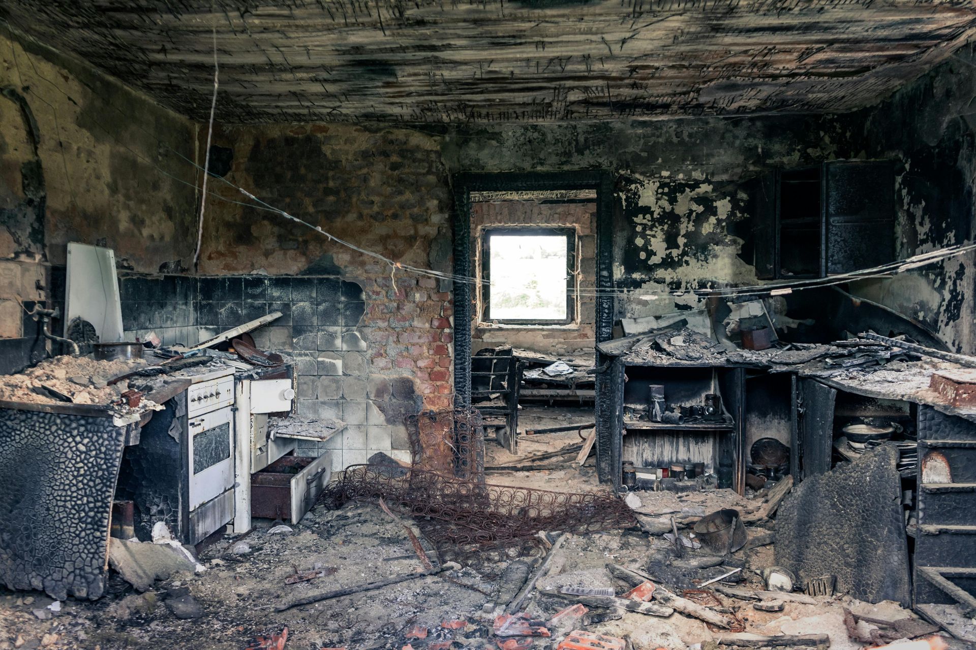 Heavily damaged kitchen interior after a fire. Walls and appliances are charred and blackened. Debris and rubble on floor.