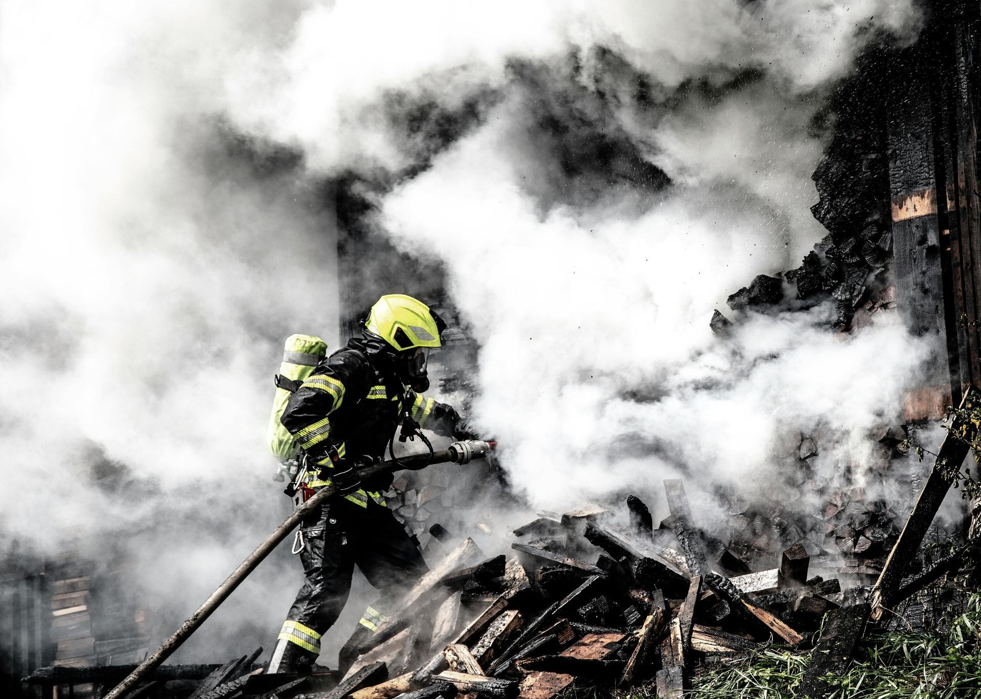 A firefighter in bright gear battles a smoky blaze, spraying water over smoldering wood debris.