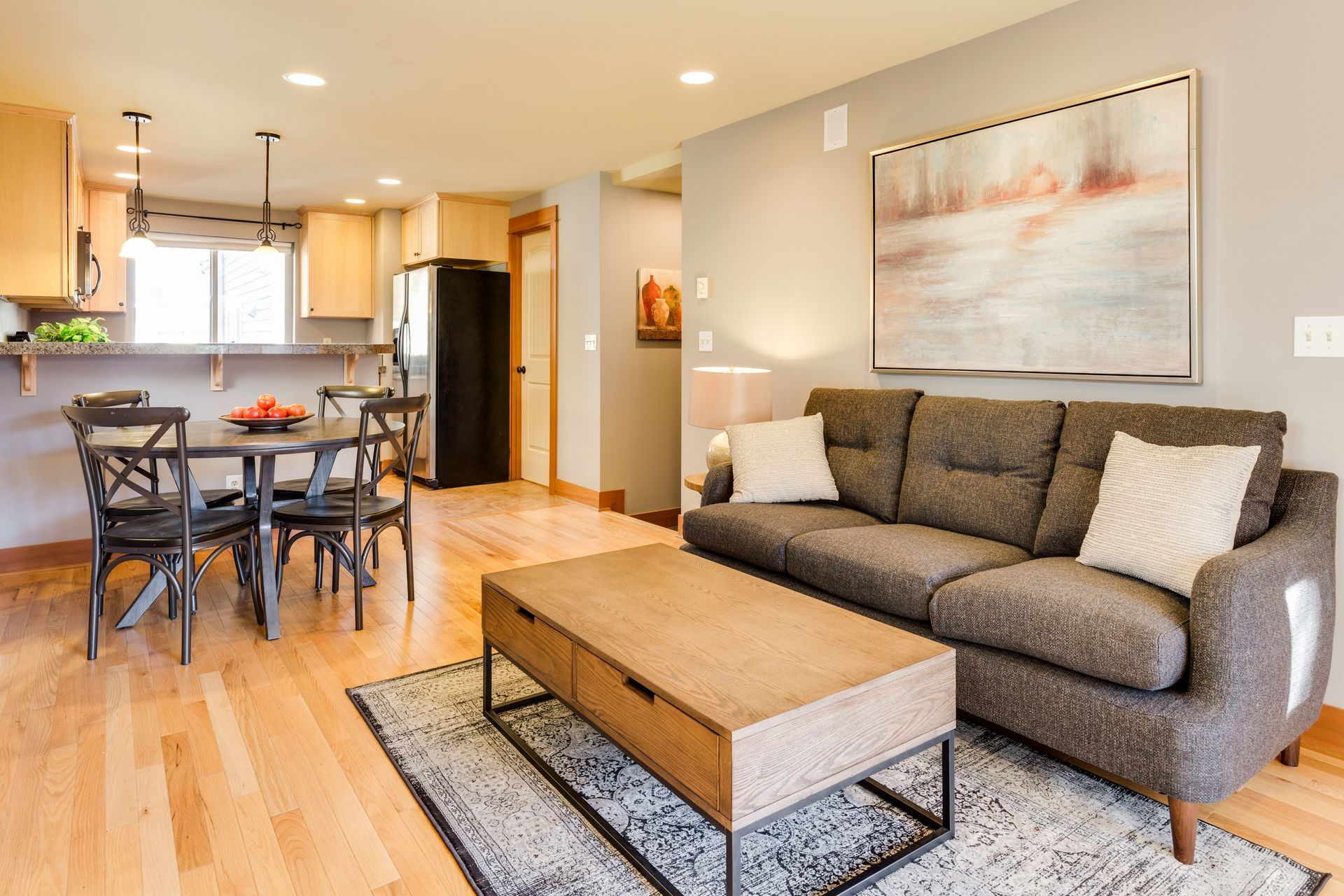 Bright open-plan living room with gray sofa, coffee table, dining set, and kitchen in the background