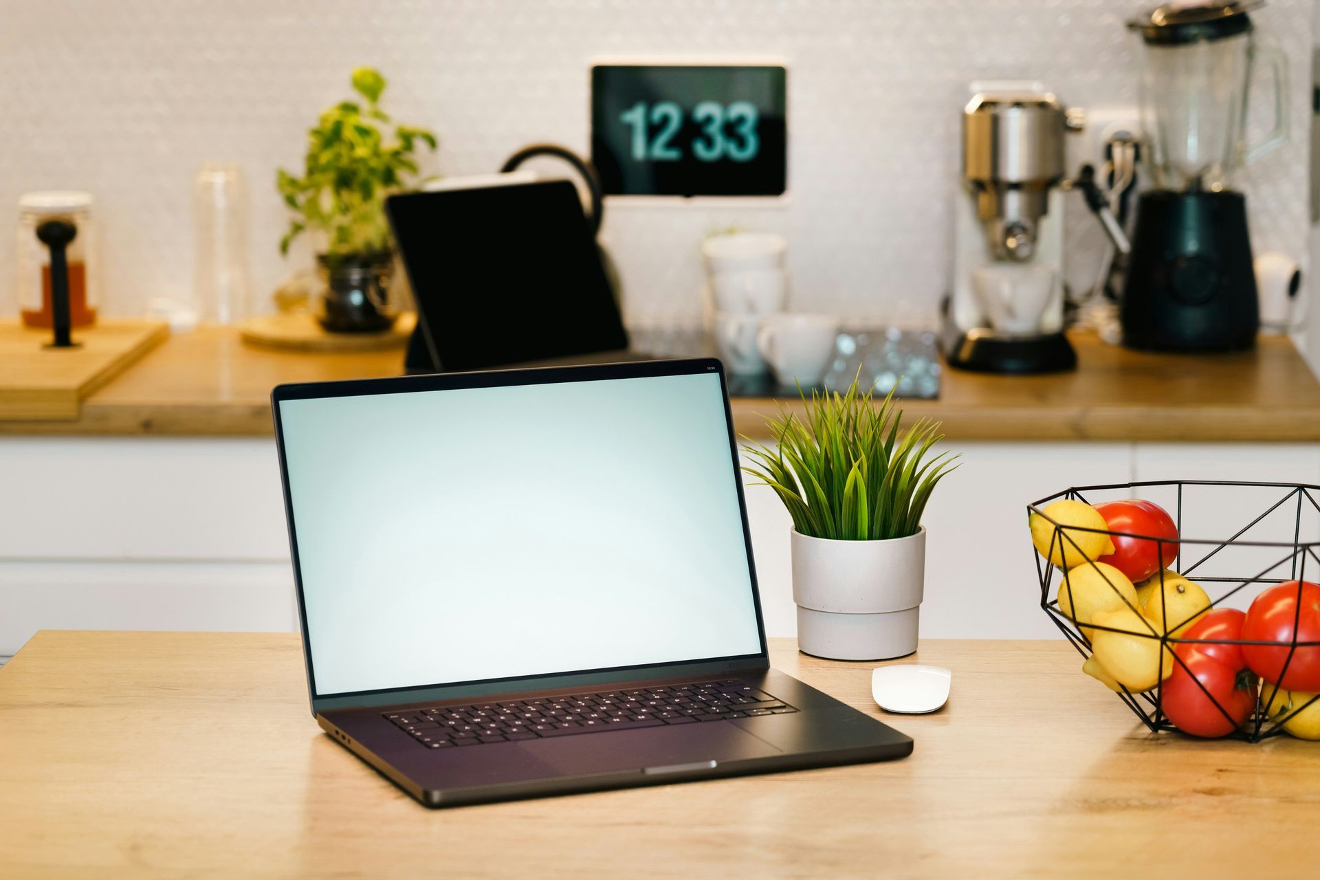 Laptop on a wooden kitchen counter with a potted plant, fruit bowl, and coffee maker in the background