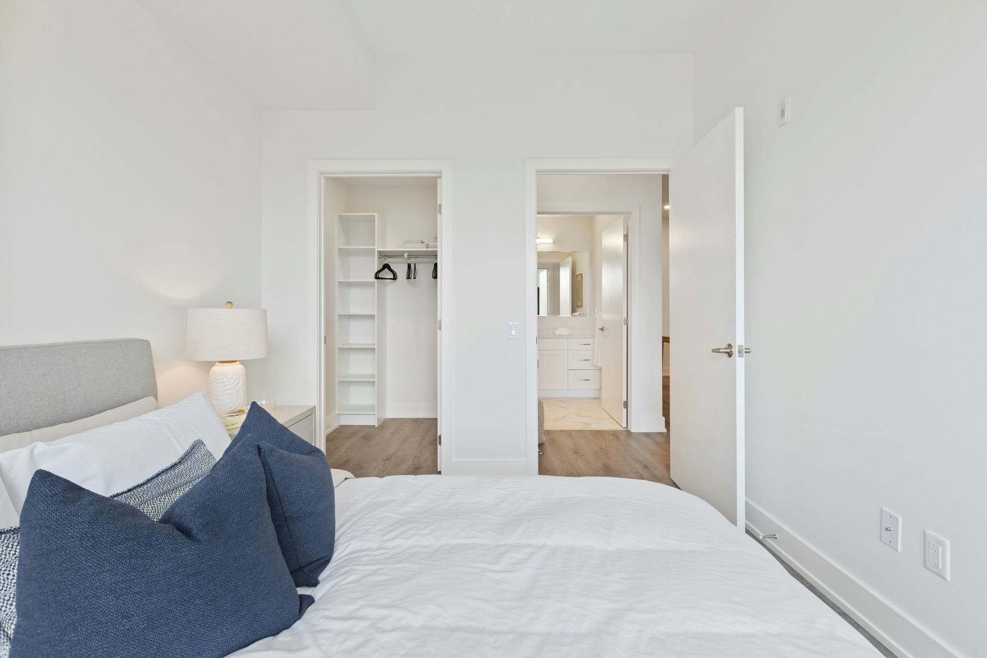 Bright white bedroom with a neatly made bed, blue pillows, and open closets leading to a hallway
