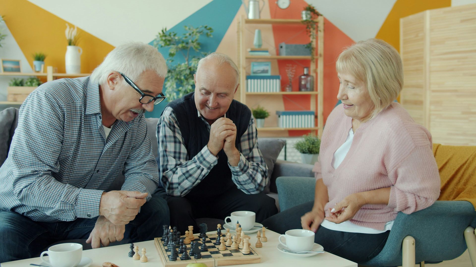 Three people play chess on a couch in a bright living room, leaning in with focused expressions.
