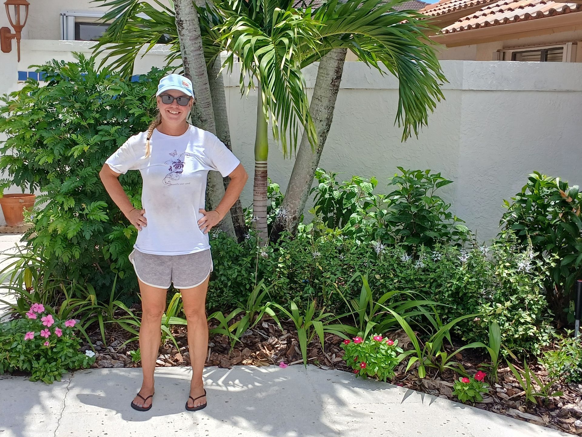 Woman in shorts and hat stands in front of lush greenery and a building with a tiled roof.