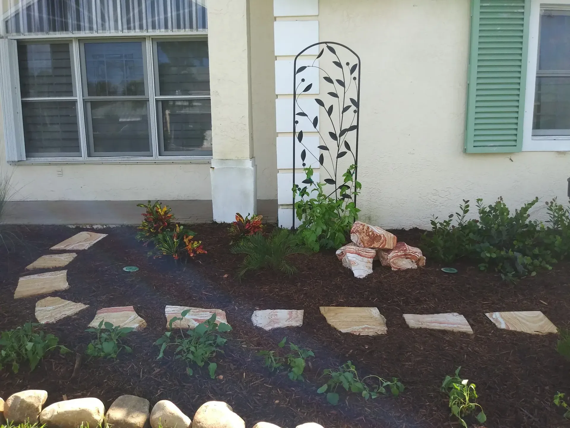 Stone path leads to a house with a green shutter and a decorative metal plant holder in a garden bed.