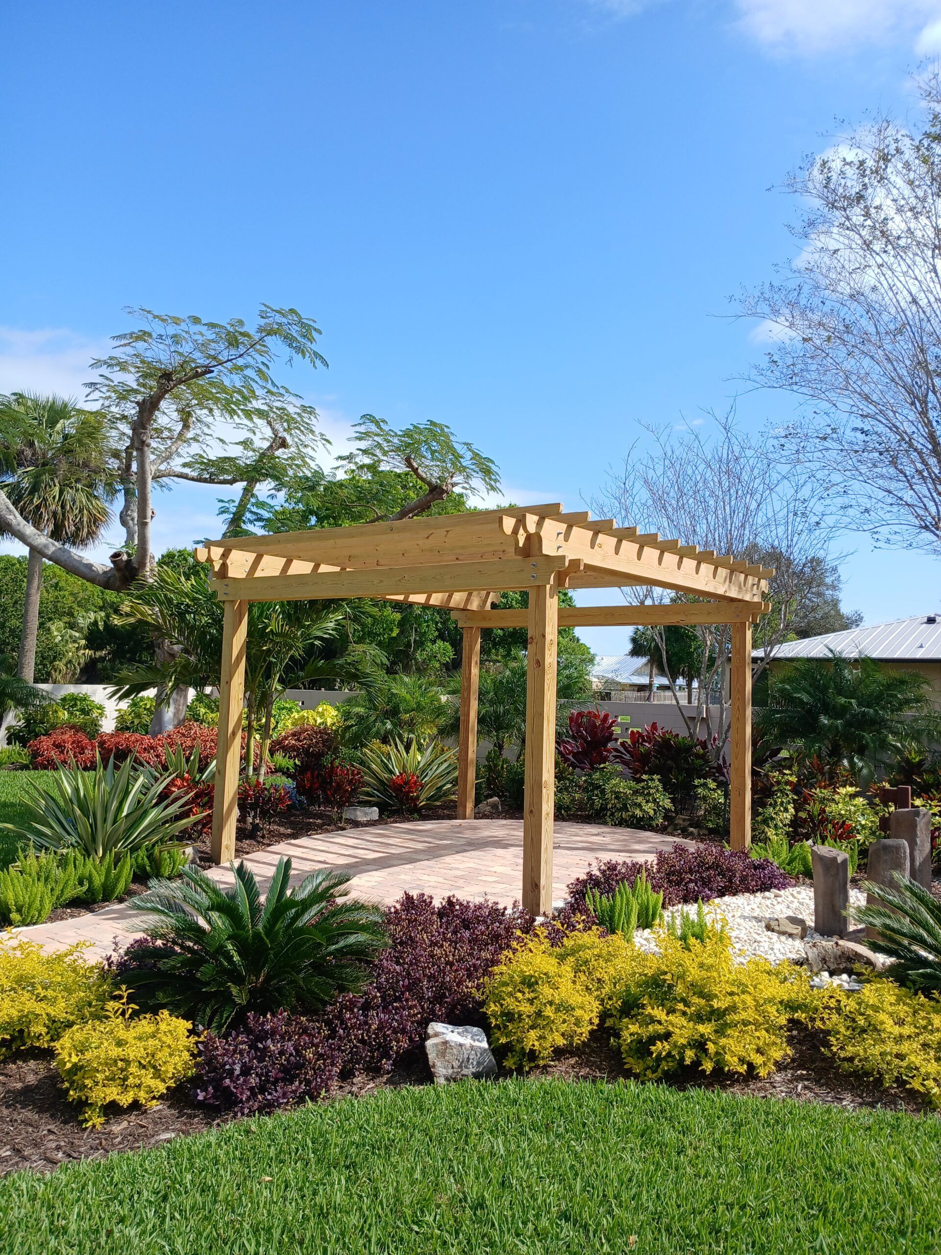 Wooden pergola in a lush garden with colorful plants and brick pathway under a blue sky.