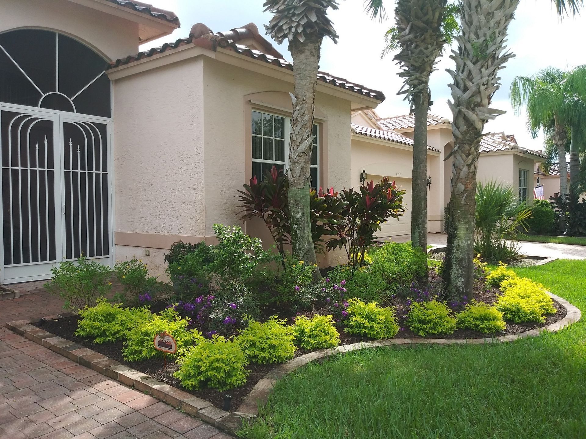 A landscaped front yard with a house, flower beds, palm trees, and green lawn.