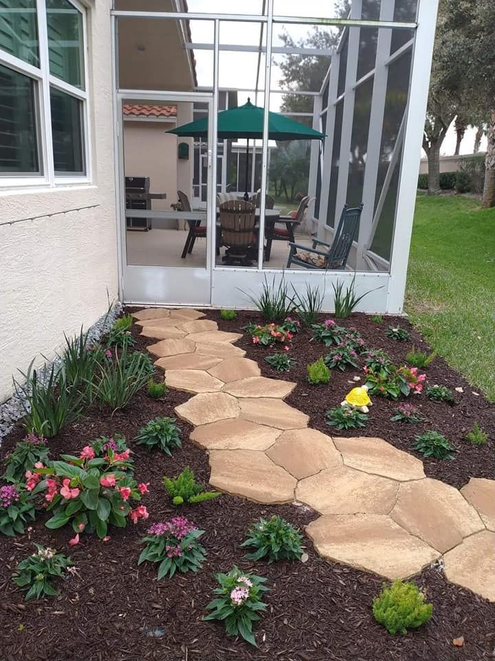 A stone pathway leads through a mulched garden with flowering plants toward a screened-in patio with outdoor furniture.