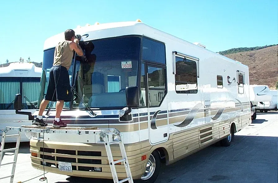 Person cleaning the windshield of a white RV in a sunny parking lot