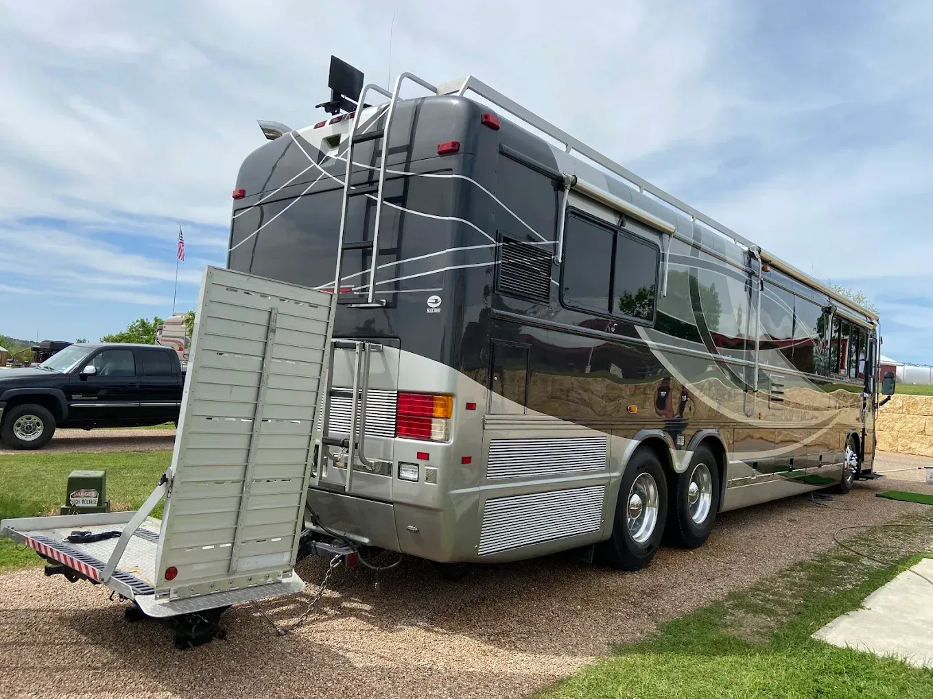Large gray RV with a rear cargo ramp parked on a gravel driveway under a cloudy sky