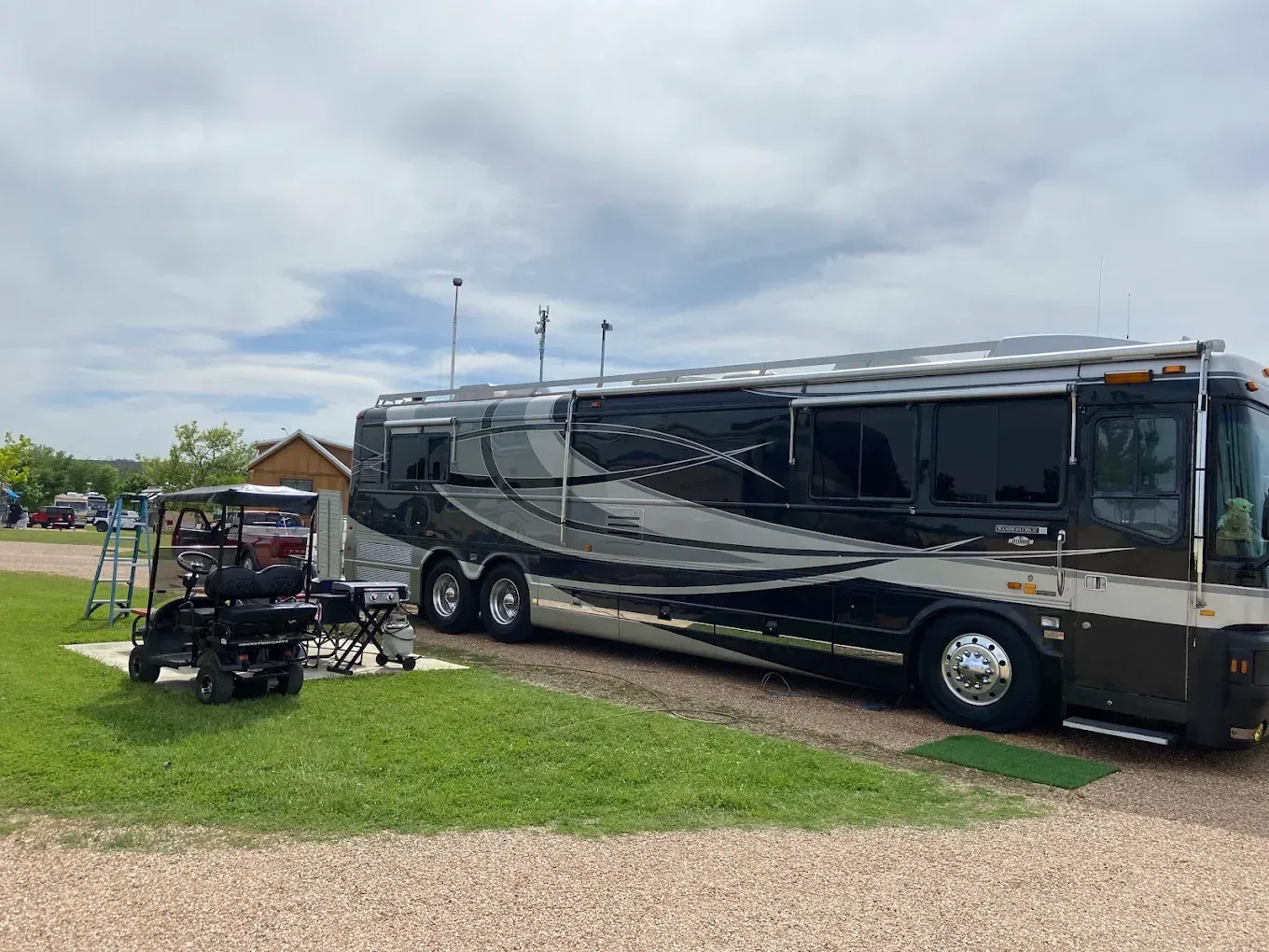 Large black-and-silver motorhome parked on grass beside a gravel lot under a cloudy sky.