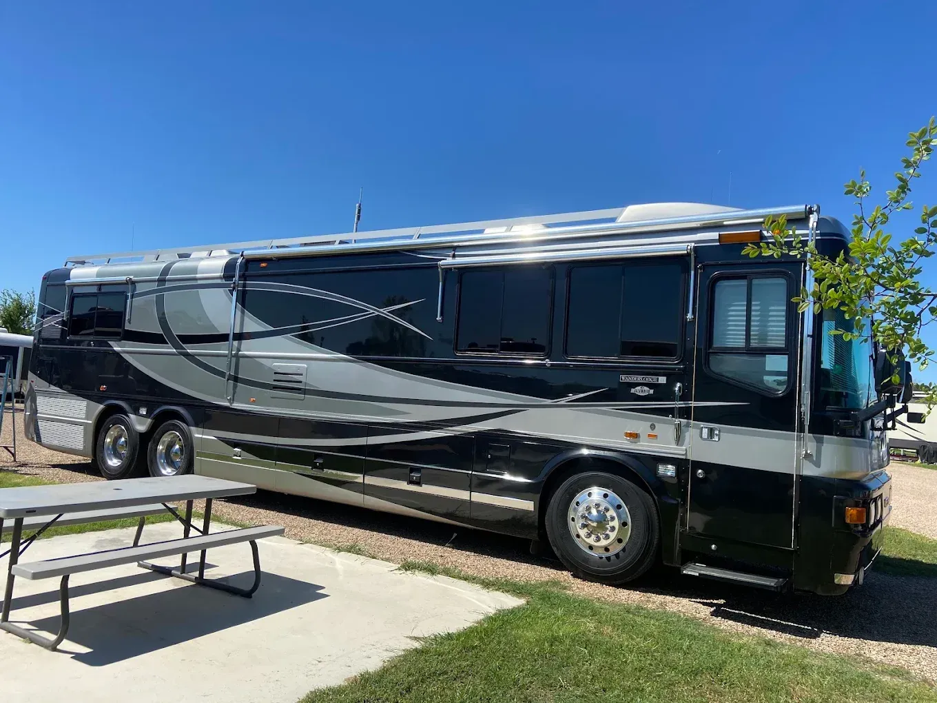 Black luxury motor coach parked in a lot under a clear blue sky, with picnic tables in the foreground