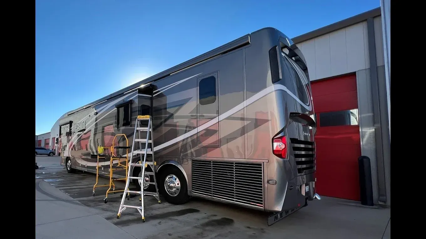 Large silver RV with ladder parked beside a building, with a red garage door and blue sky.