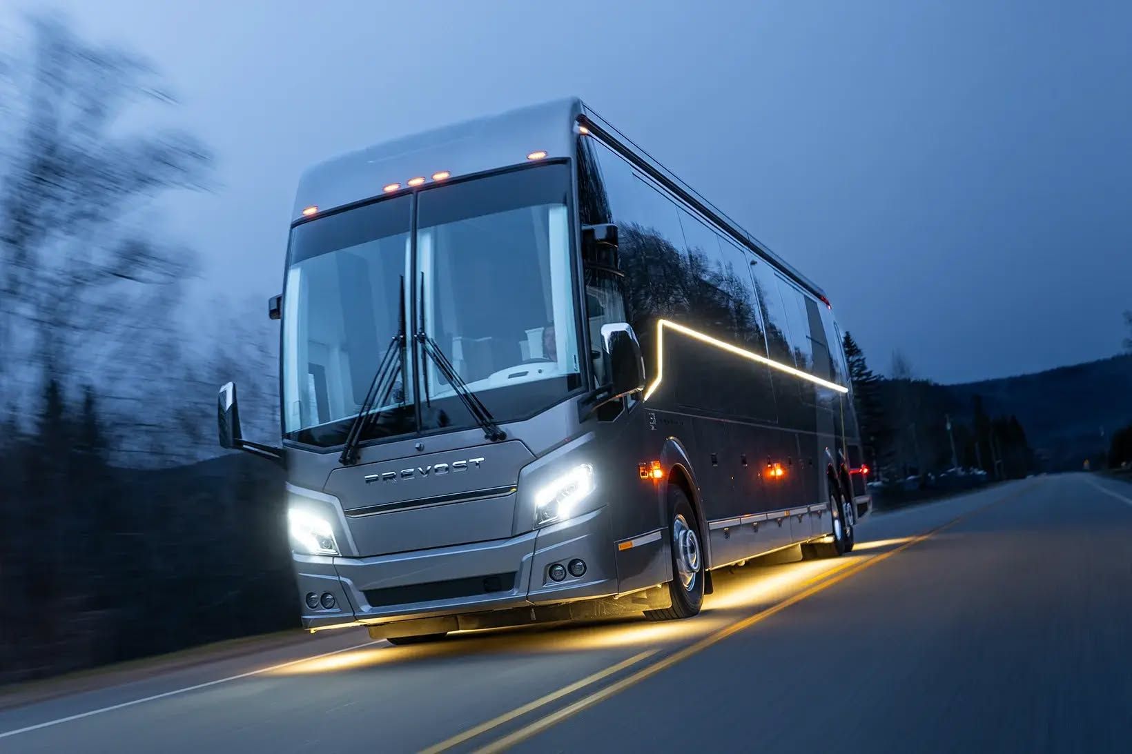 Silver coach bus driving on a dark road at dusk, headlights on, blue sky overhead