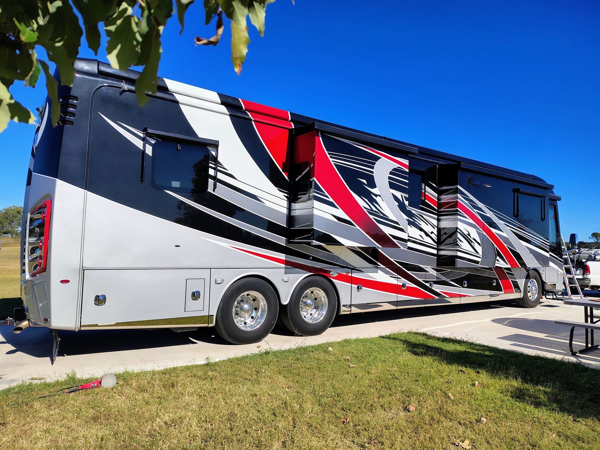 Large gray RV with red and black graphics parked beside a grassy area under a blue sky