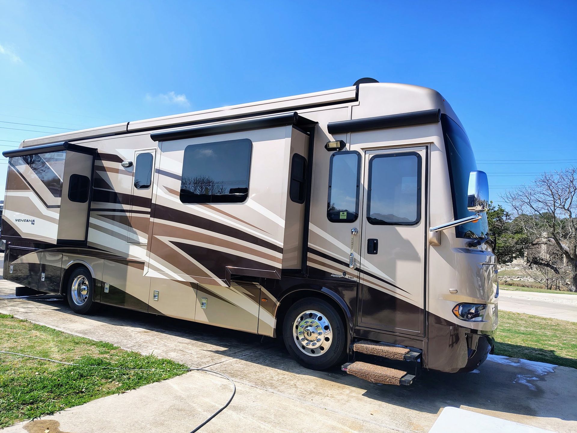 Beige and brown RV parked on a driveway beside grass under a clear blue sky.