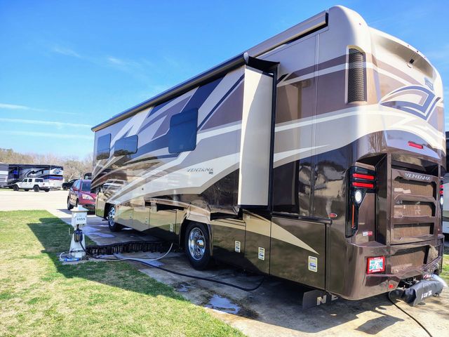 Large beige-and-brown RV parked on grass at a sunny campsite, with another RV in the background.