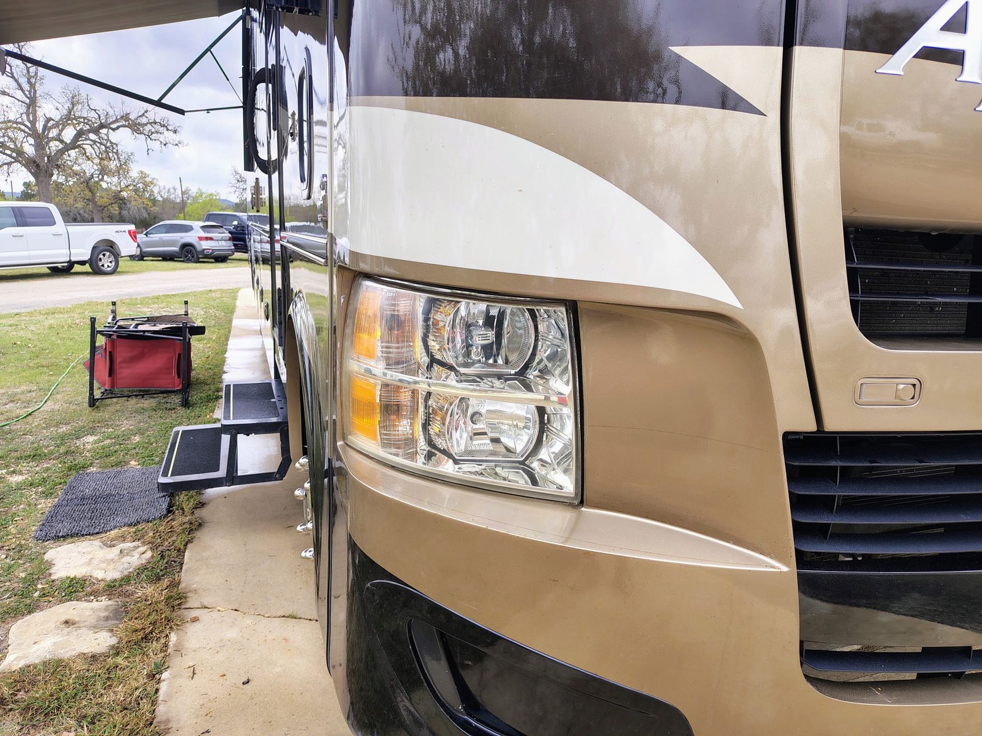 Close-up of a tan RV’s front corner and headlight at a campsite, with a table and chairs nearby.