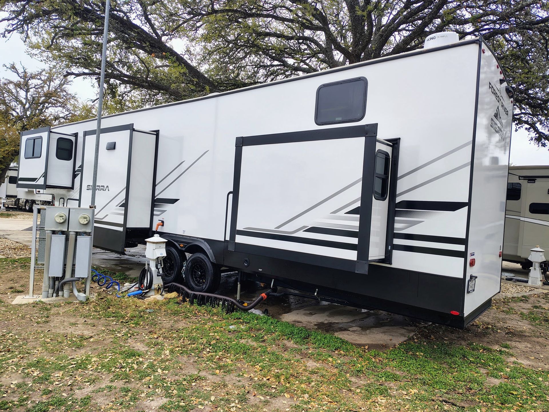 White travel trailer with awning and slide-outs parked on grass under trees