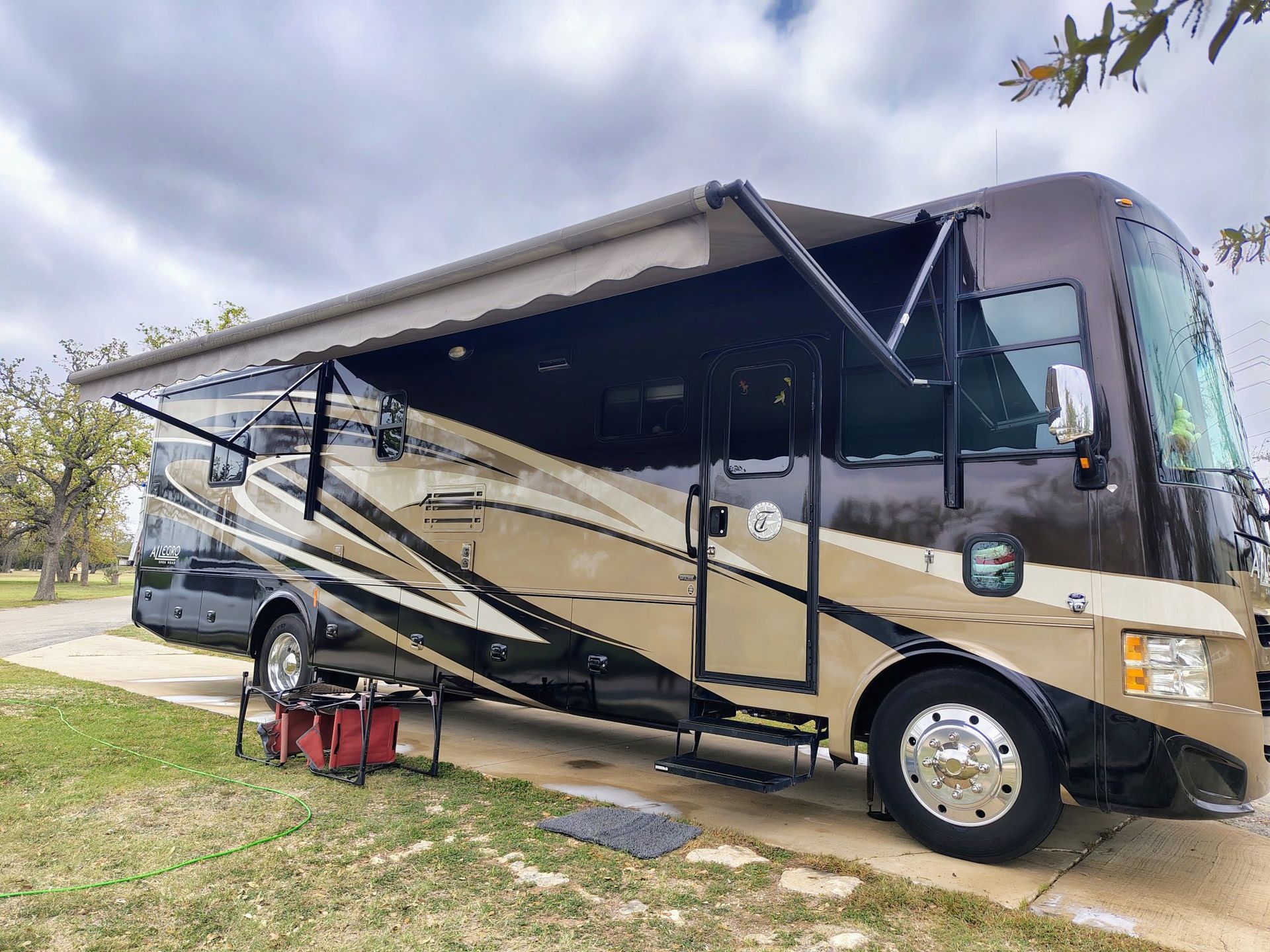 Gold and black RV with an awning extended, parked on grass at a campsite.