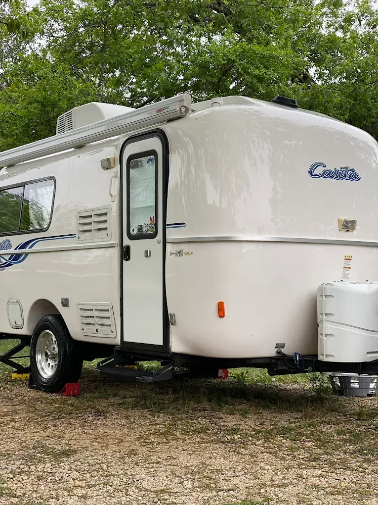 White compact travel trailer parked on gravel under trees, with blue decals and a side door.