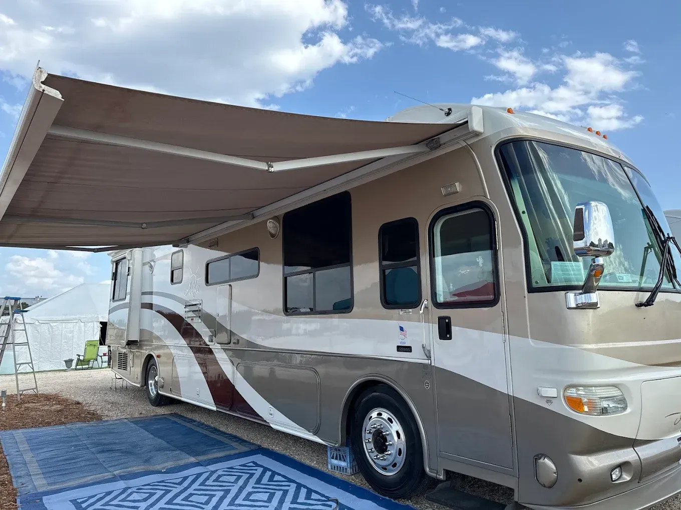 White and tan RV with an awning extended, parked on a paved lot under a blue sky