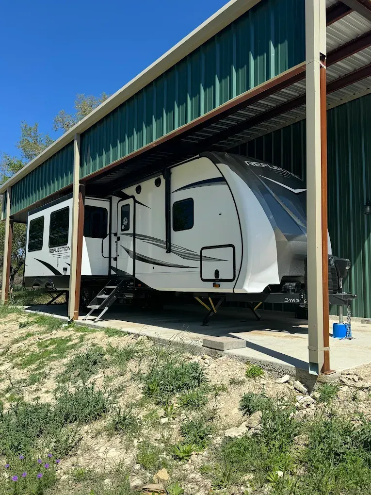 White travel trailer parked under a metal carport on a sunny day, with another RV beside it.