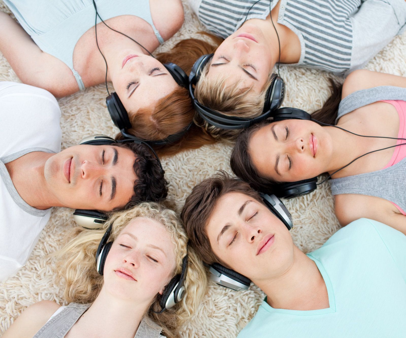 Six people lying in a circle with eyes closed, wearing headphones on a carpeted floor.