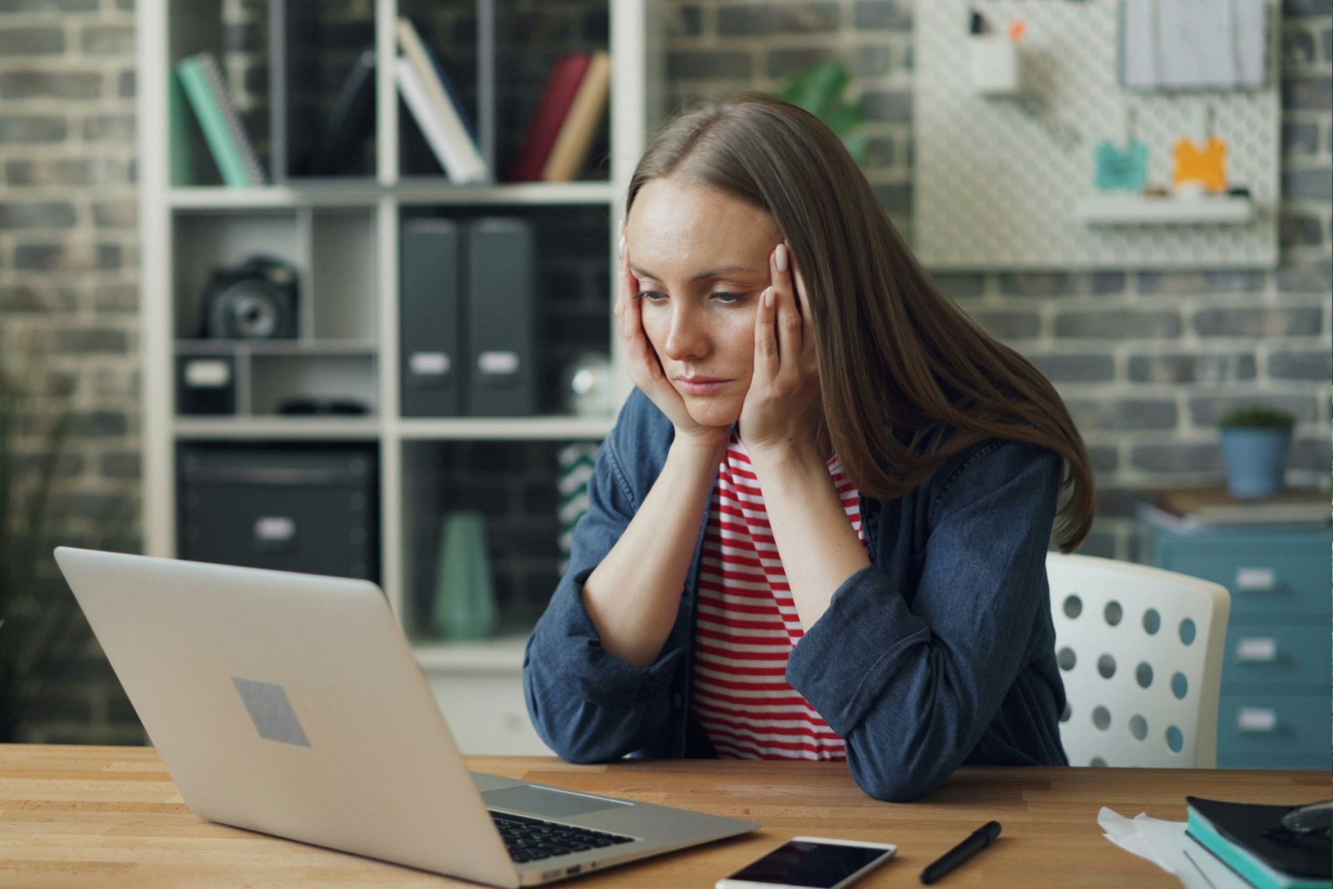 Person sitting at a desk with head in hands, looking stressed at a laptop in a home office.