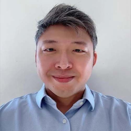 Man with gray hair, wearing a light blue button-down shirt, smiling.