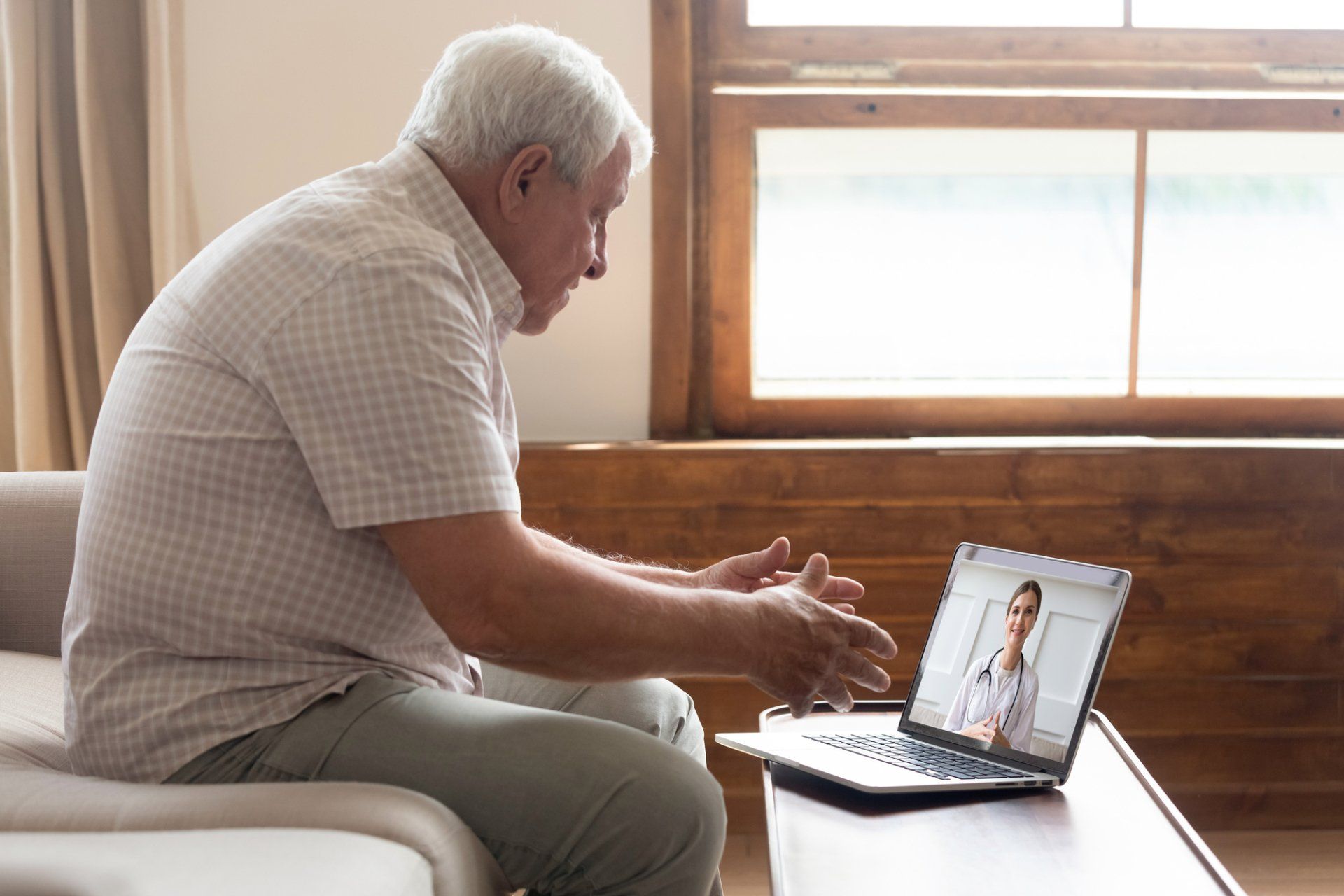 Older man in telehealth session on laptop. He sits on a couch, gestures while speaking with the doctor on screen.