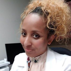 Woman with curly blonde hair, wearing a white coat and patterned top, looking at the camera.